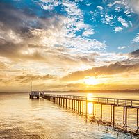 Lange Seebrücke mit weißem Geländer und kleinem Gebäude am Ende, die bei Sonnenuntergang über das ruhige Wasser führt. Goldene Sonnenstrahlen spiegeln sich auf der Wasseroberfläche unter einem wolkenreichen Himmel.