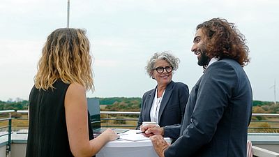 Drei Geschäftsleute stehen an einem Tisch auf einer Terrasse und unterhalten sich lachend bei einem Treffen.