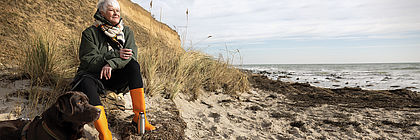 Eine Frau sitzt mit einer Thermoskanne an einer Steilküste am Strand. Neben ihr liegt ein brauner Labrador. - zum Artikel 'Die Tourismuswirtschaft in Schleswig-Holstein rückt enger zusammen – neue Winterkampagne „Küstenweisheiten“'