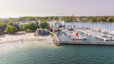Luftaufnahme des Hafens von Eckernförde mit zahlreichen Segelbooten, einer Mole mit rot-weißem Leuchtfeuer und einem angrenzenden Sandstrand. Im Hintergrund sind ein Riesenrad, die Stadt und grüne Bäume zu sehen.