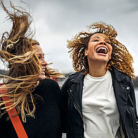 Zwei lachende Frauen gehen auf einem Steg entlang, im Hintergrund ist eine Stadt zu sehen. Die Haare der beiden Frauen wehen im Wind. 