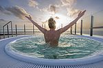 Man kan se en kvinde bagfra, som strækker armene mod himlen i et turkisblåt boblebad. I det fjerne, fra tagterrassen, hvor boblebadet er placeret, kan man se himlen, solen, et par skyer og Vesterhavet ud for Büsum i Slesvig-Holsten. 