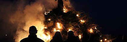 Ein großes Biikefeuer brennt. Im Vordergrund des Feuers befinden sich die Silhouetten von Menschen.  - zum Artikel 'Knisterndes Spektakel in Nordfriesland: Biikebrennen'