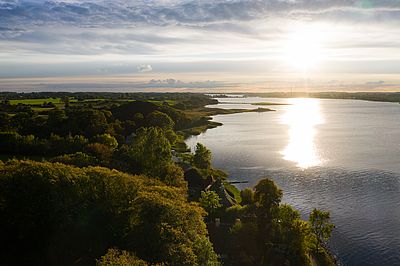 Luftaufnahme des Ostseefjords Schlei mit bewaldetem Ufer und kleinen Häusern im warmen Licht der tief stehenden Sonne.